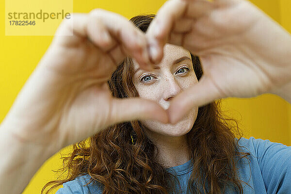 Woman gesturing heart shape in front of yellow wall
