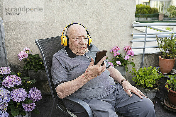 Älterer Mann hält Smartphone in der Hand und hört Musik über drahtlose Kopfhörer auf dem Balkon