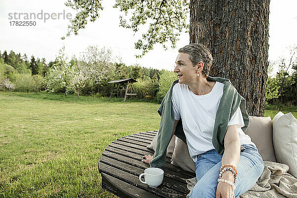 Lächelnde Frau mit Kaffeetasse sitzt auf Bank vor Baumstamm im Garten