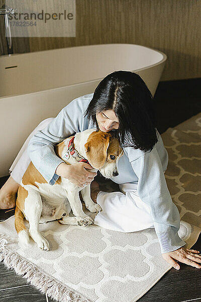Frau umarmt Hund  der auf Teppich neben Badewanne im Badezimmer sitzt