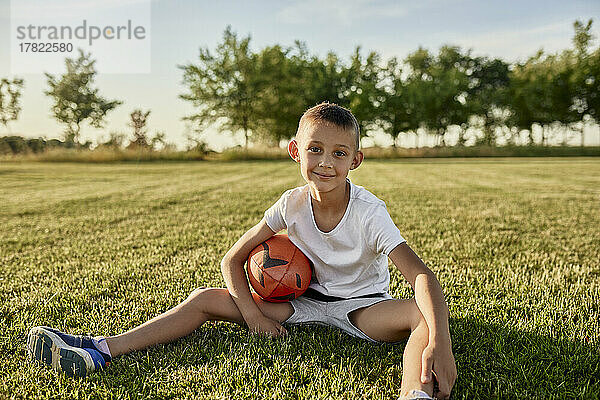 Lächelnder Junge mit Rugbyball sitzt an einem sonnigen Tag auf dem Sportplatz