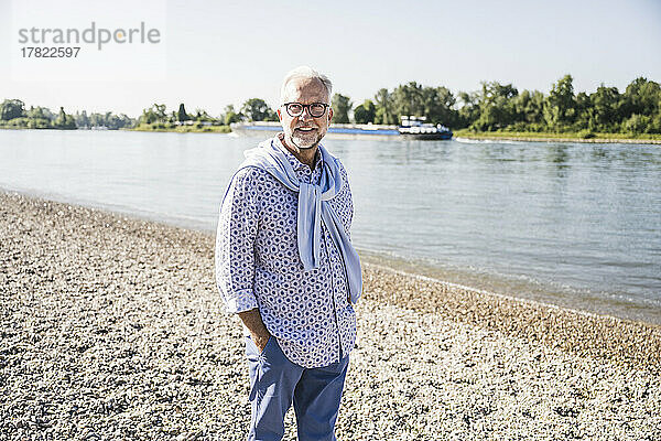 Lächelnder älterer Mann steht mit der Hand in der Tasche am Flussufer