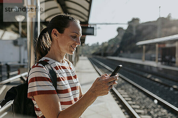 Lächelnde Frau  die am Bahnhof steht und ihr Smartphone benutzt