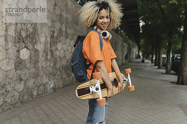 Lächelnde Frau mit Rucksack hält Skateboard auf Fußweg