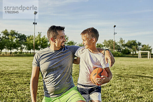 Glücklicher Vater mit Arm um den Sohn  der an einem sonnigen Tag Rugbyball hält