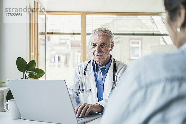 Leitender Arzt benutzt Laptop vor Patient in medizinischer Klinik