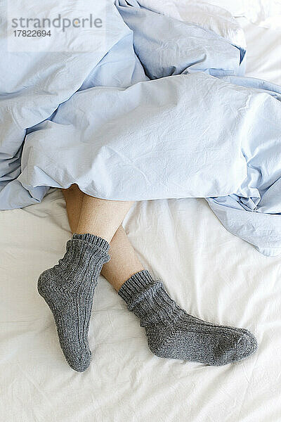 Legs of woman wearing cozy socks under blanket on bed