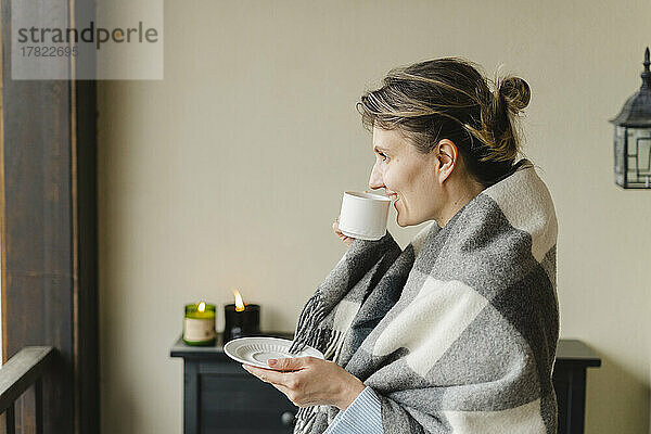 Lächelnde Frau  in eine Decke gehüllt  trinkt zu Hause Kaffee