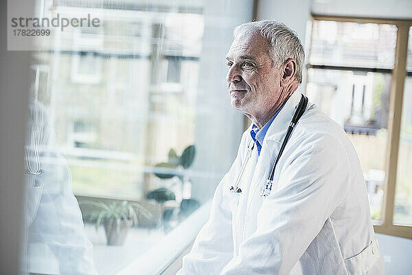 Lächelnder Arzt  der durch das Fenster auf das Krankenhaus blickt