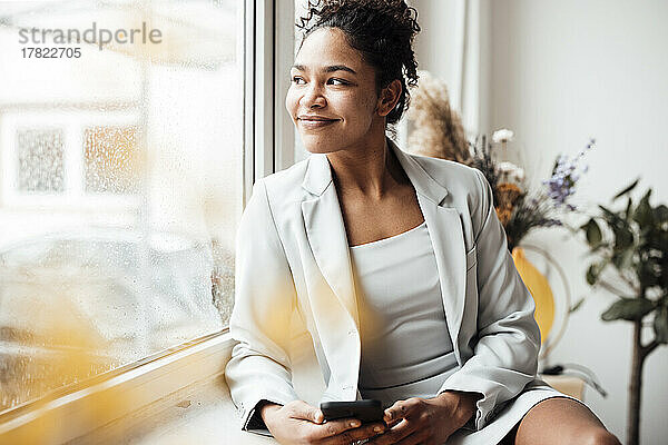 Lächelnde Geschäftsfrau mit Smartphone  die durch das Fenster schaut