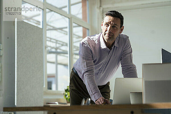 Nachdenklicher Geschäftsmann mit Laptop  der sich im Büro auf den Tisch lehnt