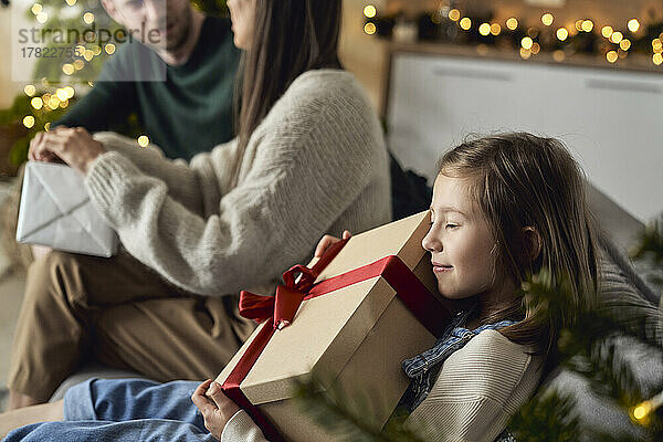 Lächelndes Mädchen umarmt Geschenkbox  die zu Hause bei den Eltern sitzt