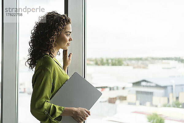 Geschäftsfrau hält Laptop und schaut aus dem Fenster im Büro