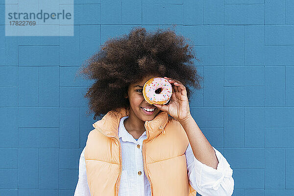 Lächelndes Mädchen mit Afro-Frisur  das das Auge mit Donut vor blauer Wand bedeckt