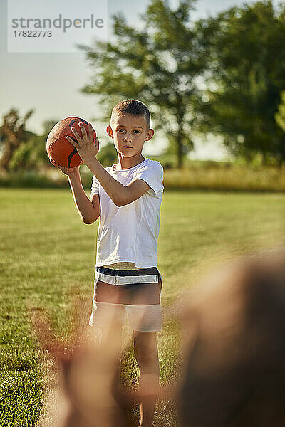 Junge hält Rugbyball und steht an einem sonnigen Tag auf dem Sportplatz