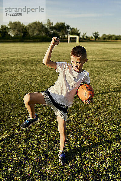 Junge mit Rugbyball macht Haka-Tanz und steht auf einem Bein auf dem Sportplatz