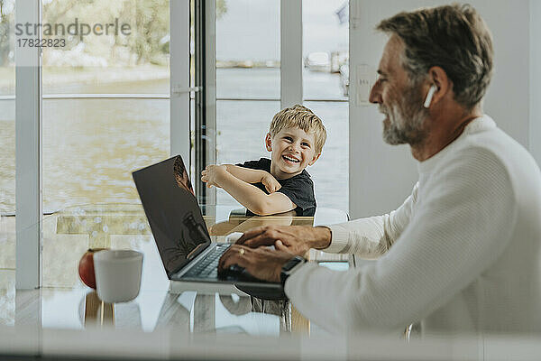 Fröhlicher Junge schaut Vater an  der am Laptop arbeitet