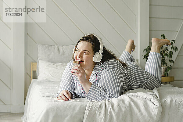Glückliche Frau isst Schokolade und hört Musik über kabellose Kopfhörer  die zu Hause im Bett liegt