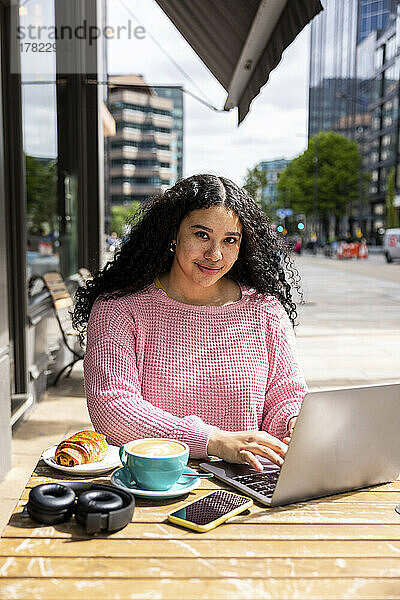 Lächelnde junge Frau mit Laptop sitzt an einem sonnigen Tag im Straßencafé