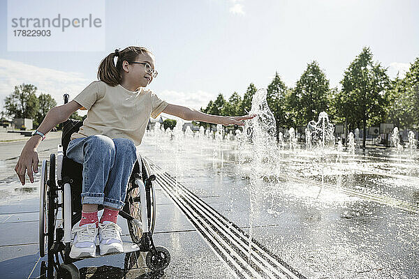 Mädchen mit Behinderung berührt an sonnigem Tag spritzendes Brunnenwasser im Park