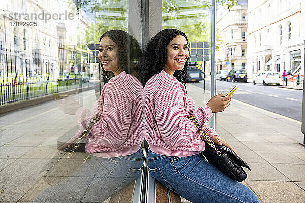 Glückliche junge Frau mit Smartphone sitzt auf Glasfenster