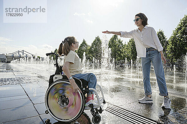 Mädchen mit Behinderung blickt Mutter zu  die an einem sonnigen Tag mit spritzendem Brunnenwasser spielt