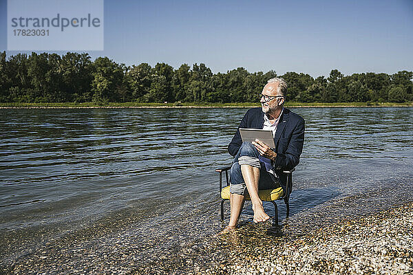 Mann mit Tablet-PC sitzt auf Stuhl am Flussufer