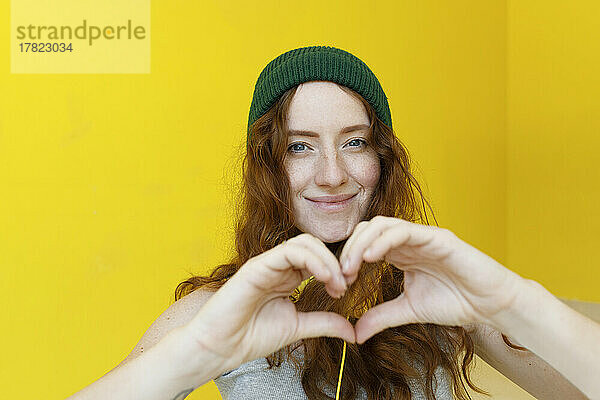 Smiling woman gesturing heart shape in front of yellow wall