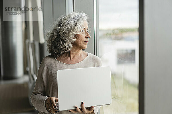 Ältere Geschäftsfrau mit Laptop schaut aus dem Fenster im Büro