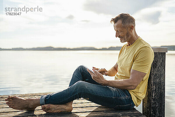Reifer Mann sitzt mit Tablet-PC am Pier am See