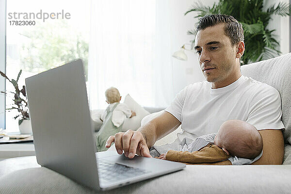 Mann trägt Baby im Arm und arbeitet von zu Hause aus am Laptop