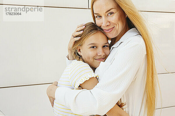 Glückliche Frau mit blonden Haaren umarmt Tochter vor der Wand