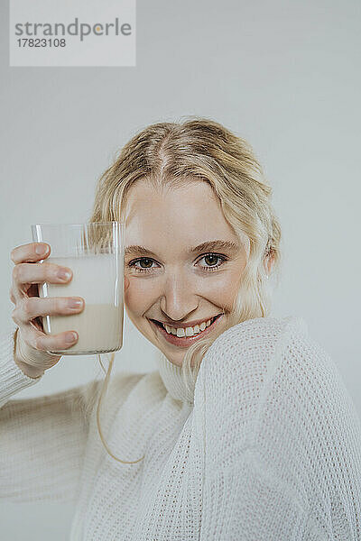 Lächelnde Frau hält ein Glas Pflanzenmilch vor weißem Hintergrund