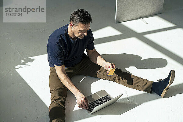 Geschäftsmann benutzt Laptop und hält Kreditkarte in der Hand  während er im Büro auf dem Boden sitzt