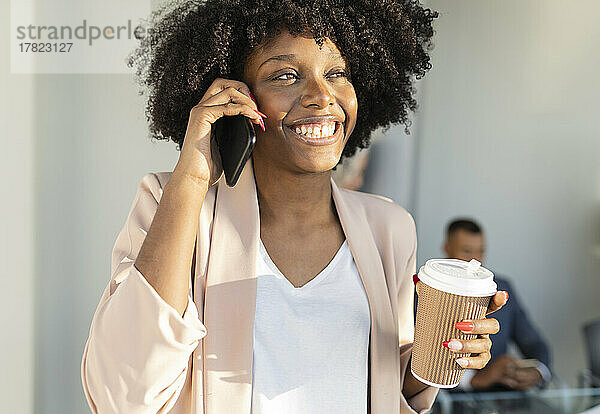 Glückliche Geschäftsfrau  die am Smartphone spricht und einen Einwegbecher in der Hand hält