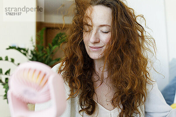 Frau mit zerzausten Haaren hält kleinen tragbaren Ventilator zu Hause