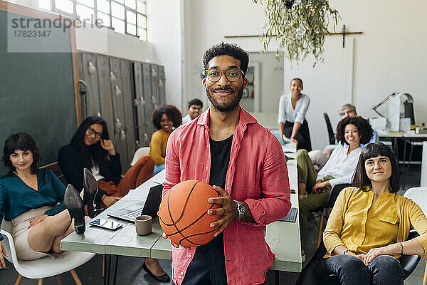 Lächelnder Geschäftsmann mit Basketball von multiethnischen Kollegen im Büro