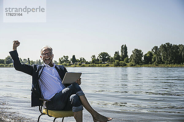 Glücklicher Mann hält Tablet-PC in der Hand und gestikuliert am Flussufer