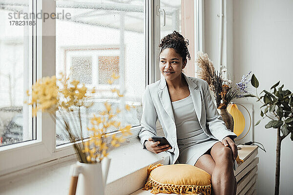 Lächelnde Geschäftsfrau mit Smartphone sitzt am Fenster