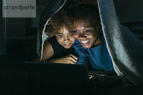 Lächelnde Frau zeigt ihrer Tochter zu Hause unter der Decke einen Tablet-PC