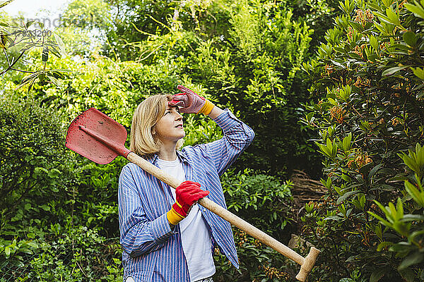 Frau schirmt Augen ab und trägt Schaufel in der Nähe von Pflanzen im Garten