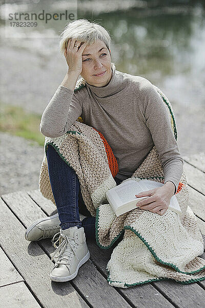 Reife Frau mit Buch und Decke sitzt auf dem Pier