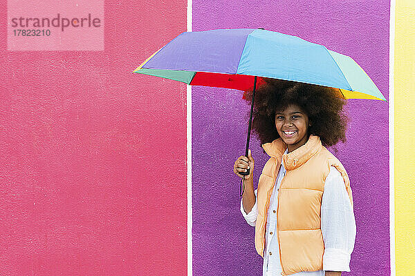 Smiling girl holding multi color umbrella in front of wall