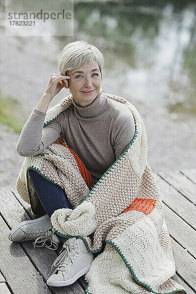 Lächelnde Frau sitzt mit Strickdecke am Pier