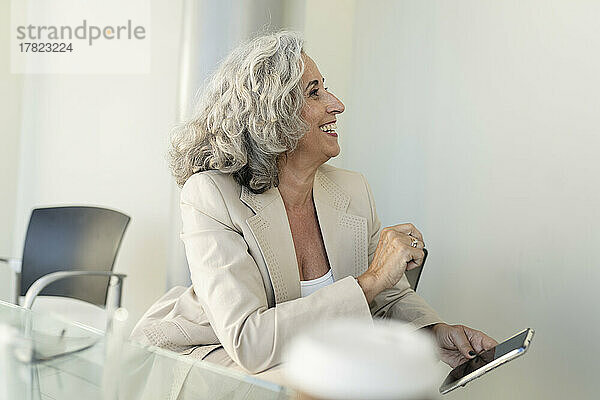 Lächelnde ältere Geschäftsfrau mit Smartphone sitzt im Büro