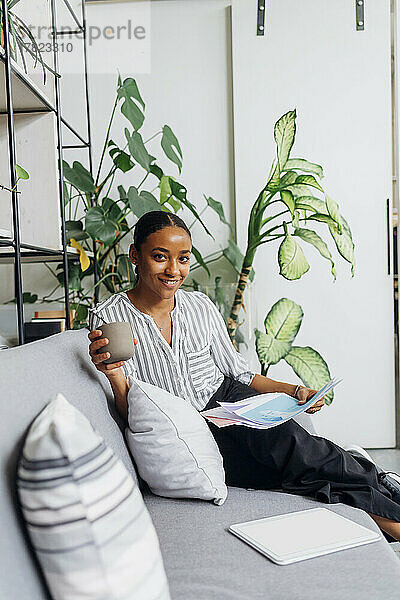 Lächelnde Geschäftsfrau mit Dokumenten und Kaffeetasse auf dem Sofa im Büro