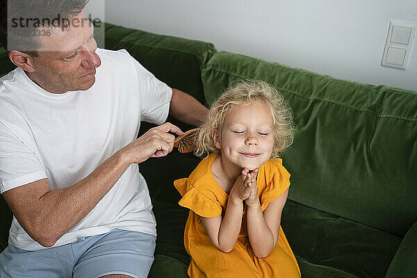 Lächelndes Mädchen mit geschlossenen Augen sitzt auf dem Sofa  während ihr Vater sich zu Hause die Haare kämmt