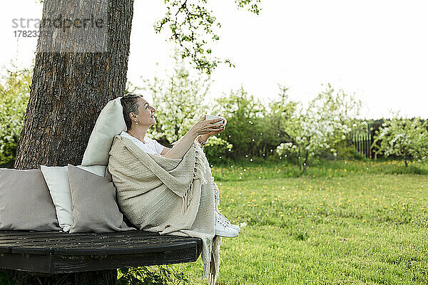 Glückliche Frau  in eine Decke gehüllt  hält eine Kaffeetasse in der Hand und sitzt auf einer Bank um einen Baumstamm