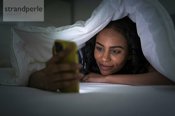 Junge Frau unter einer Decke bedeckt und benutzt Smartphone im Schlafzimmer