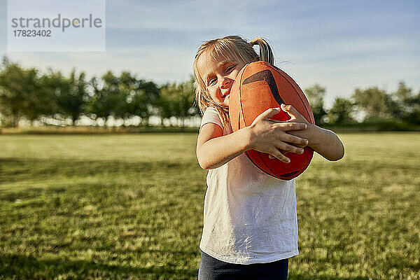 Lächelndes Mädchen umarmt Rugbyball auf dem Sportplatz an einem sonnigen Tag